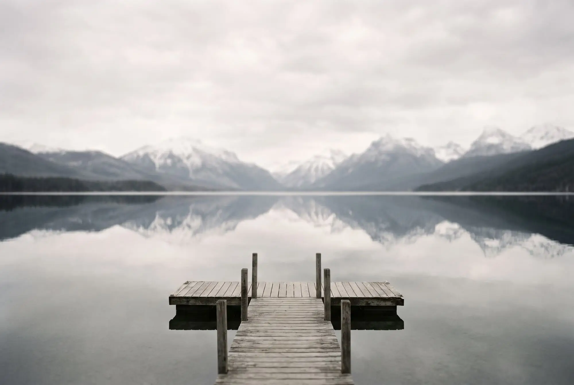 A still alpine lake with a wooden jetty, snowy peaks mirrored in the water