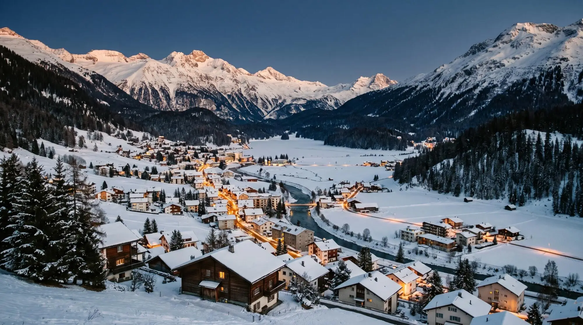 A snowy mountain town in a wide alpine valley at dusk, with warm lights along the river
